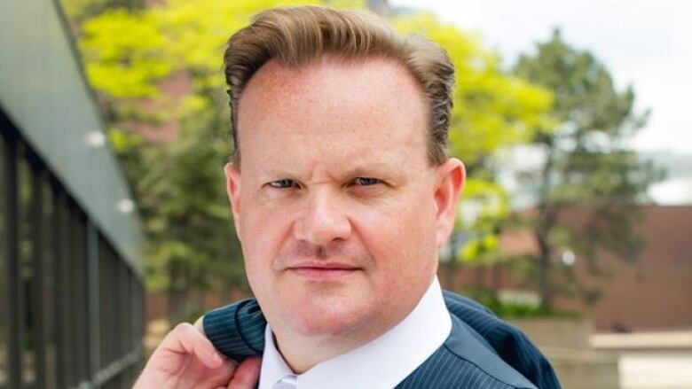 A man in a suit holds his blazer over his shoulder and stares intensely at the camera, brow somewhat furrowed.