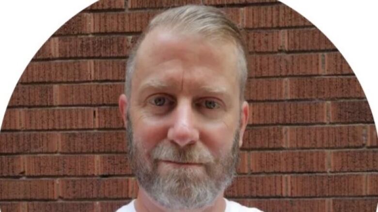 A man with a grey beard stares at the camera from in front of a red brick wall.