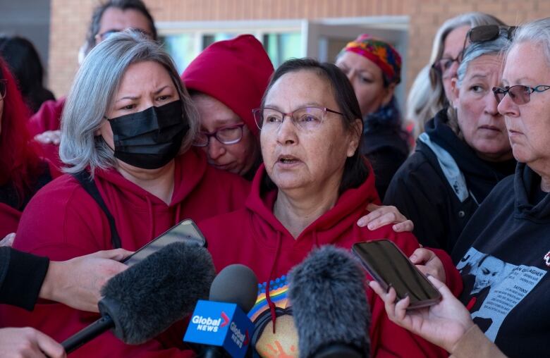 A woman wearing a red sweater and glasses stands in front of several microphones and recording devices. She is surrounding by other women who are holding onto her. 