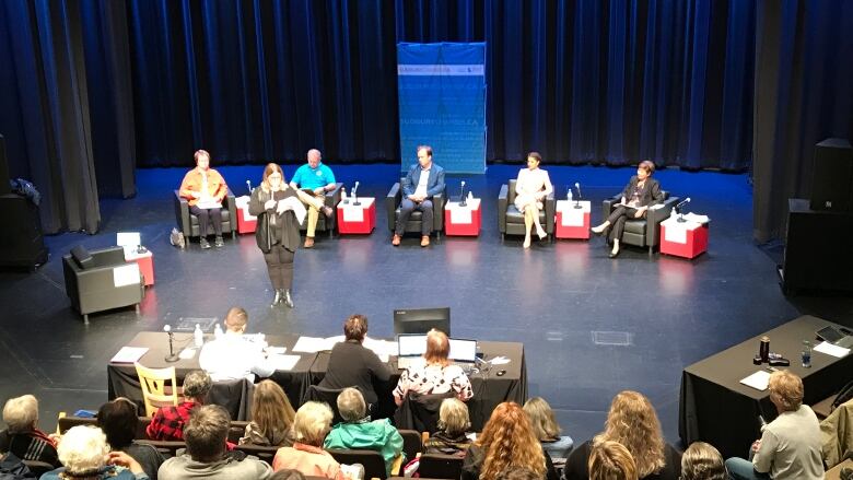 People sit in chairs on a stage preparing for a debate.