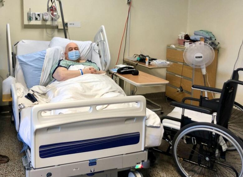 A bald, masked man in his hospital bed next to a wheelchair.