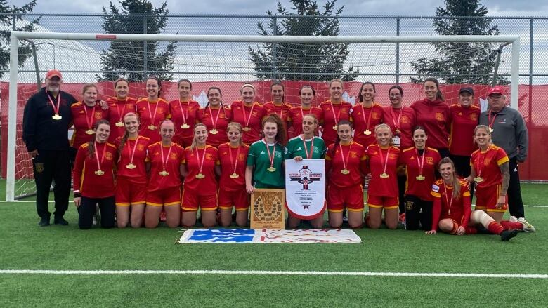 Two rows of soccer players kneel and stand in front of a soccer net with a plaque and a Newfoundland flag in front of them.