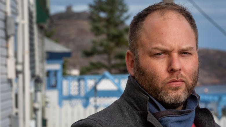 A man wearing a grey jacket looks into the camera. He has short hair and a short beard.