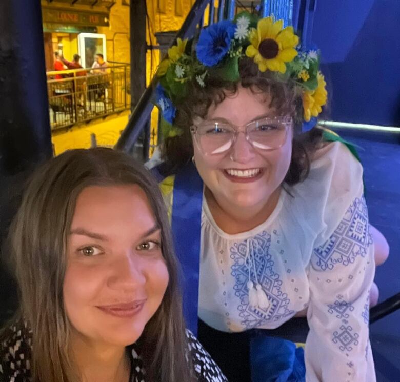 Two women smile for a photograph. One has long brown hair, while the other has shorter curly hair and is wearing a blue and white outfit and a floral headdress.