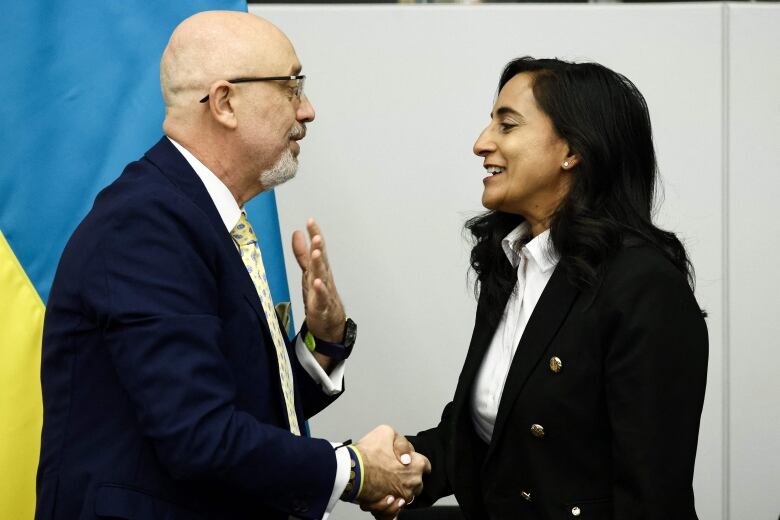 A man and a woman shake hands in front of a Ukrainian flag.