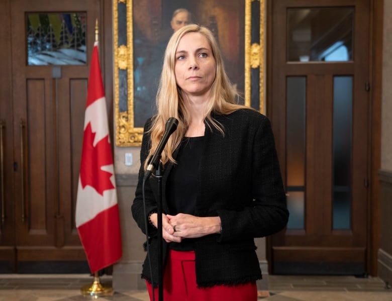 A blonde woman wearing a black top and blazer stands in front of a microphone with a Canadian flag in the background.