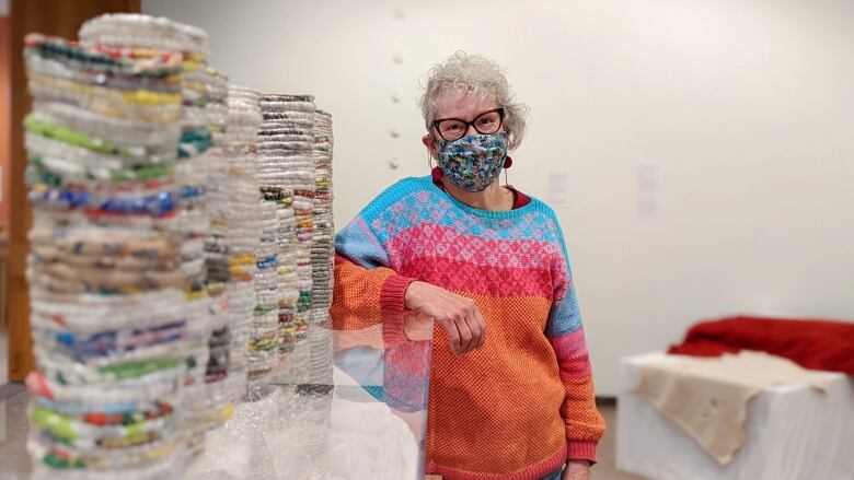 A woman wearing a brightly coloured knit sweater leans on an artist plinth holding plastic sculptures.