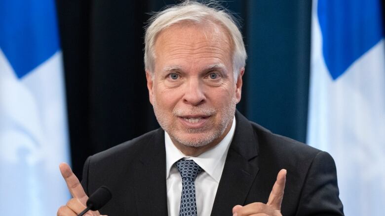 A man wearing a suit is speaking while seated in front of Quebec flags.