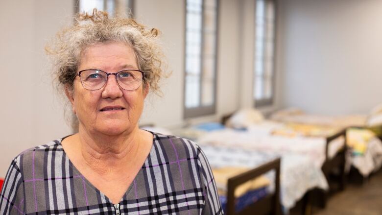 An older woman with glasses stands in front of emergency shelter beds.