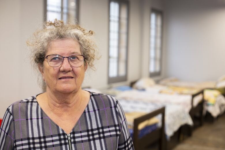 An older woman with glasses stands in front of emergency shelter beds.