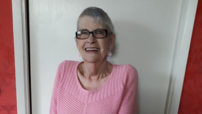 A woman with short grey hair, glasses and a pink knitted top stands smiling with crutches. 