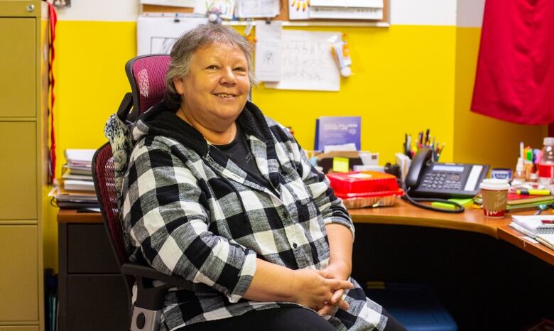 An older woman with short hair wearing a plaid jacket sits in her office chair surrounded by Indigenous art.