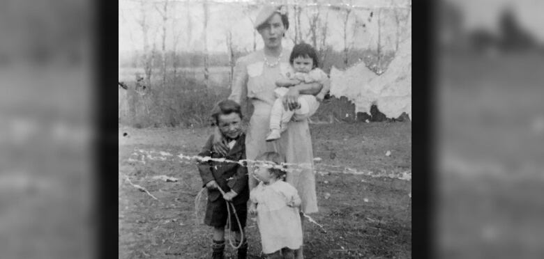 Aged photo of a woman standing, holding a baby, with two children at her feet.