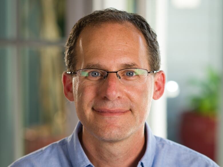 A portrait of a middle-aged gray-haired man with glasses, looking straight at the camera and smiling, wearing a light blue button-downs shirt.