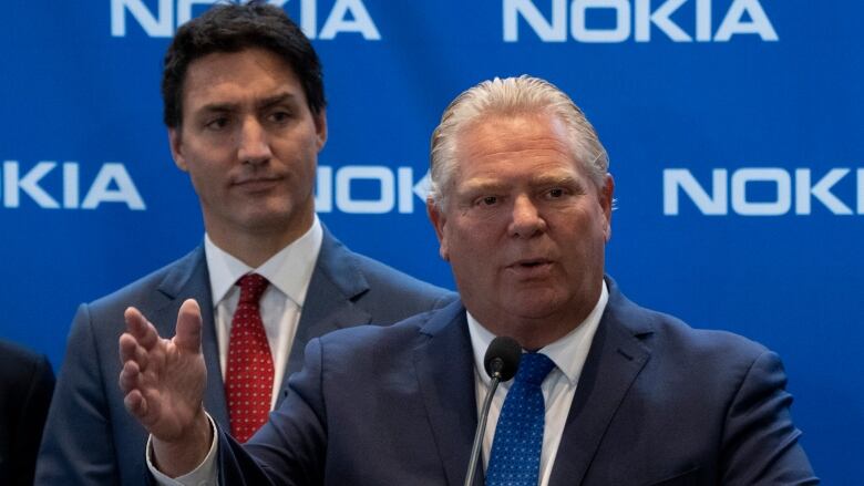 A politician speaks at a lectern in front of a blue corporate backdrop while another listens behind him.