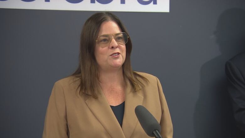 A woman with brown hair and glasses speaks into a microphone.