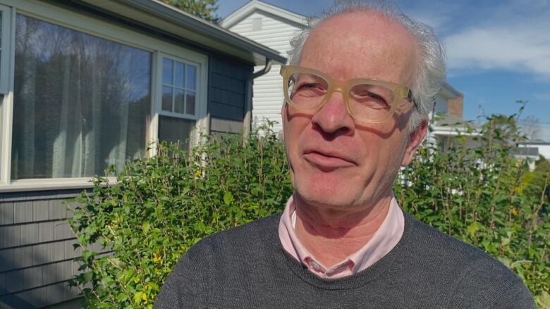 A man is seen with grey hair, wearing glasses and a dark grey sweater over a pink collared shirt.