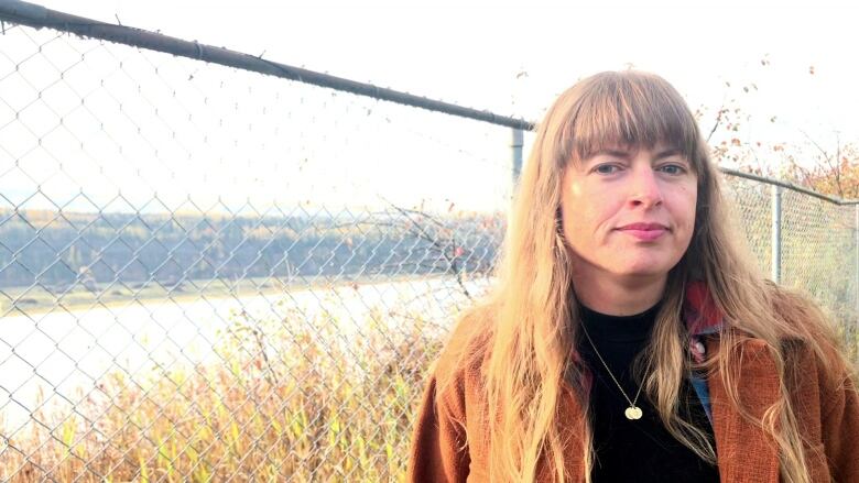 A woman in a brown jacket stands by a chainlink fence with a river running behind it.