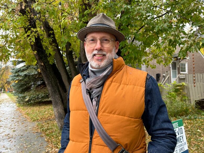 A man wearing glasses, an orange vest, blue shirt and a hat is smiling at the camera while standing on a sidewalk.