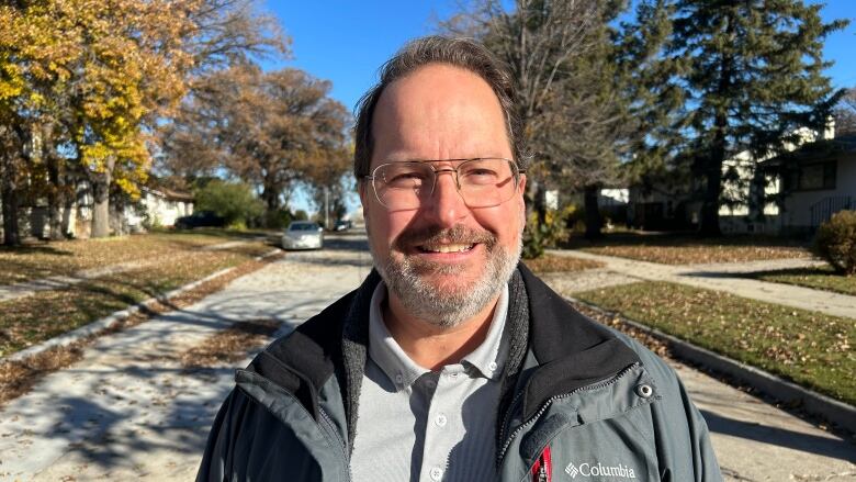 A man wearing glasses, jacket and grey shirt is standing on a road and smiling at the camera.