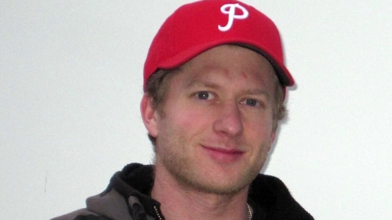 A young white man with a red baseball cap and grey and black patterned hoodie is giving a slight smile in front of a white background.