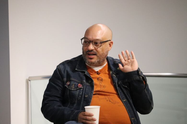 A Black man in an orange t-shirt holds up his hand. 