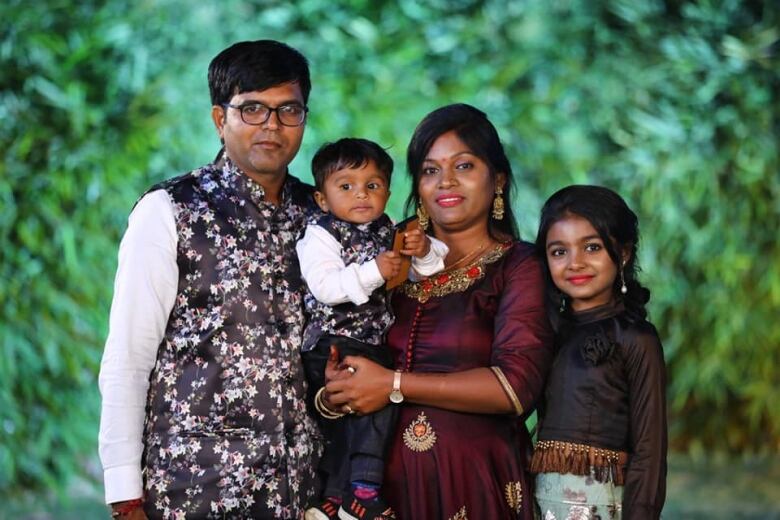 An Indian famiy of four is pictured against a green leafy backdrop. A man stands on the left hand side, a woman holds a toddler boy, and an older girl stands on her right.