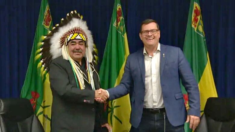 Two men stand shake hands in front of a few Saskatchewan flags at an indoor announcement