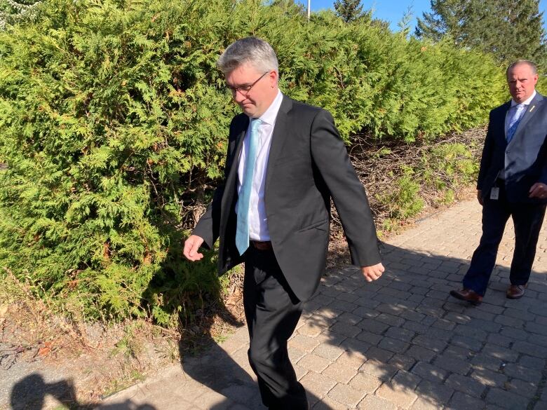 A man wearing a grey suit and powder-blue tie walks next to a green hedge.