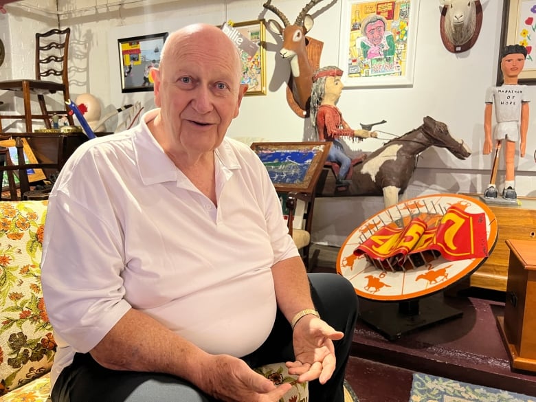 A man sits on a couch, looking at the camera. There are statues and artworks behind him.