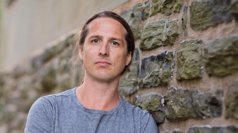 A white man with his hair in a ponytail standing against a stone brick wall. 