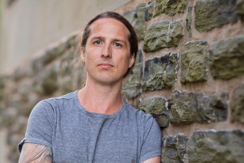 A white man with his hair in a ponytail standing against a stone brick wall. 