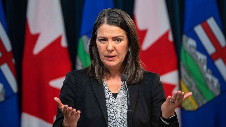 Alberta Premier Danielle Smith gestures with both hands, while standing in front of flags of Alberta and Canada.