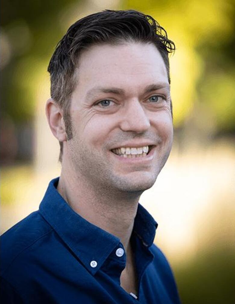 A man with short hair wearing a blue button-up shirt smiles.