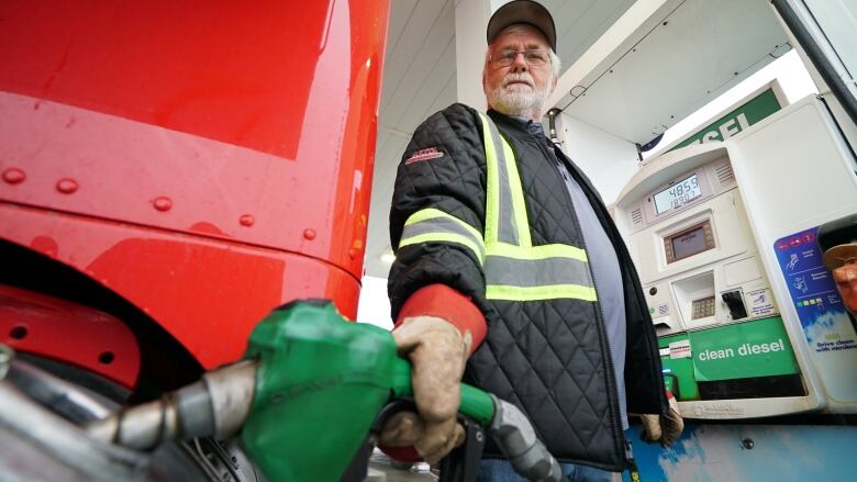 A man stands at a gas pump fueling a red transport truck. The nozzle is sitting in the fuel tank pumping diesel.