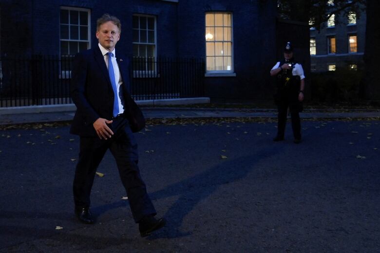 An individual wearing a blue tie and suit walks outside a London residence at night. 