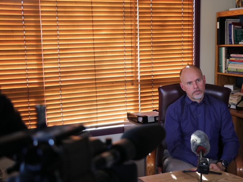 A balding man with a goatee sits at a desk in an office in front of a video camera and a microphone. 