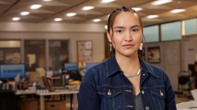 A young, Indigenous woman stands in a newsroom with desks and computers visible behind her. She wears a silver necklace and long silver earrings, her hair is tied back in a ponytail and she is only visible from the waist up wearing a denim jacket open over a grey shirt.