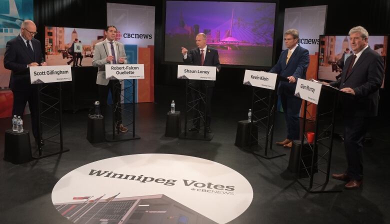 Five men are standing behind podiums on a television set. A sign on the floor says 