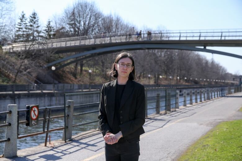 A woman with dark brown hair and glasses stands in front of the Rideau Canal. She's wearing a blazer and jeans. 