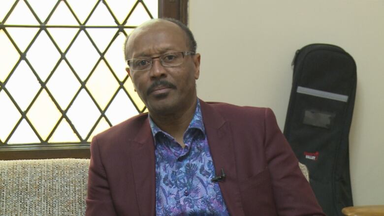 Renel Sanon sits in a maroon blazer and blue shirt with glasses inside the church. 