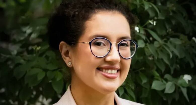 A person smiles with their face tilted slightly to the side. They are wearing glasses and are in front of a bush.