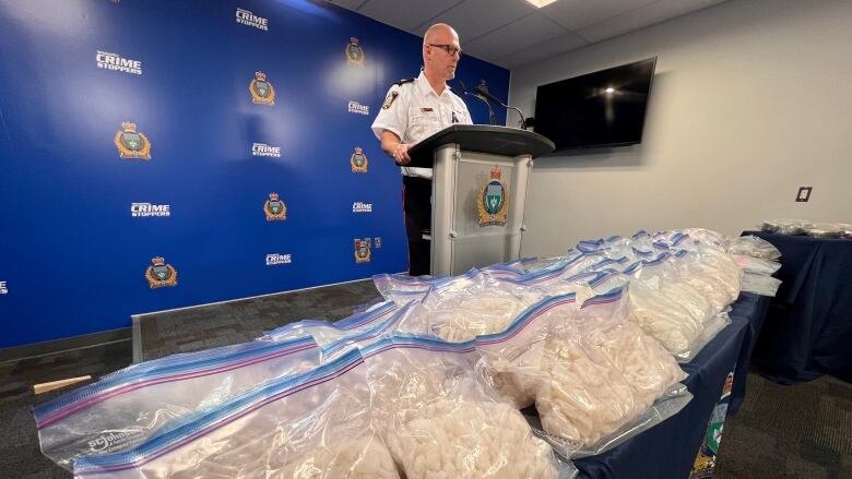 A police officer stands at a podium speaking in front of a table covered with large plastic bags filled with methamphetamine.