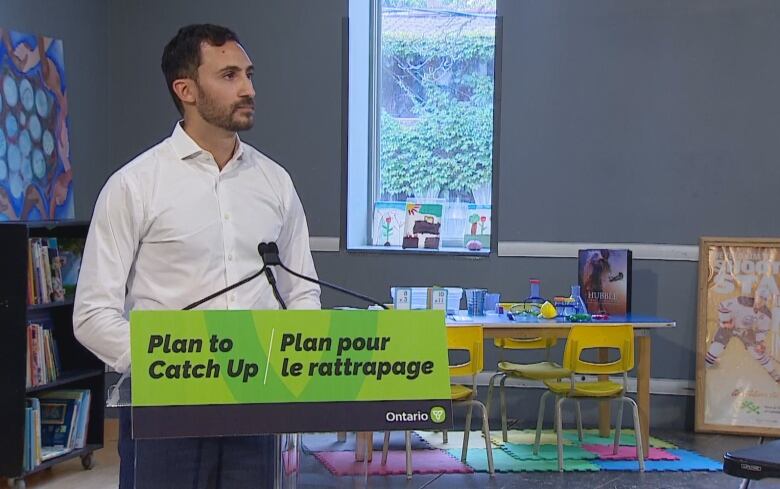 A man in a white button-up shirt stands behind a podium in an empty classroom.