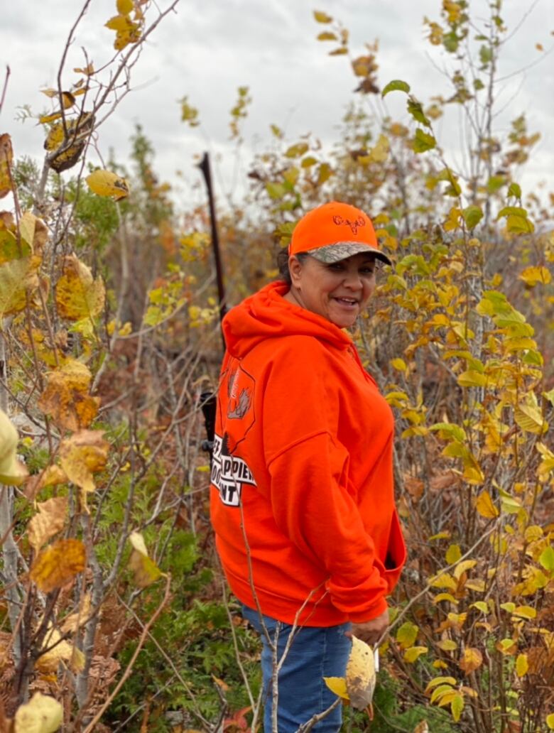 A woman in an orange shirt and baseball cap stands in some country brush. She smiles at the camera.