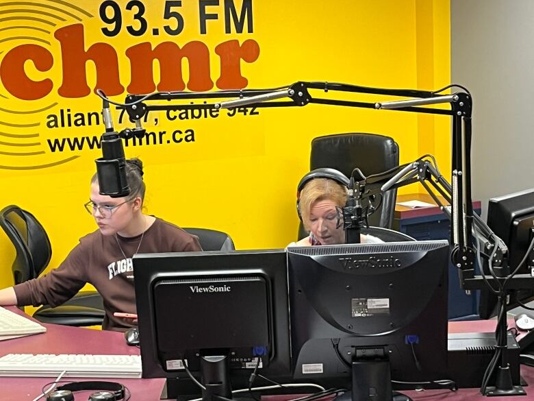 A woman wearing over the ear headphones speaks into a microphone for a radio show. Another woman sits to her left, and is handling the microphone levels and music on a computer.