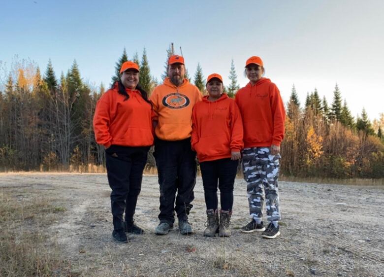 One man and three women in orange sweaters stand together in front of a forest.