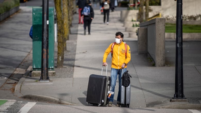 A man wearing a yellow hoodie is seen with a facemask and luggage.