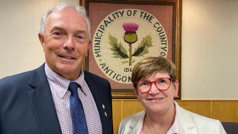 A man and a woman stand together and smile at the camera. A sign in the background says 