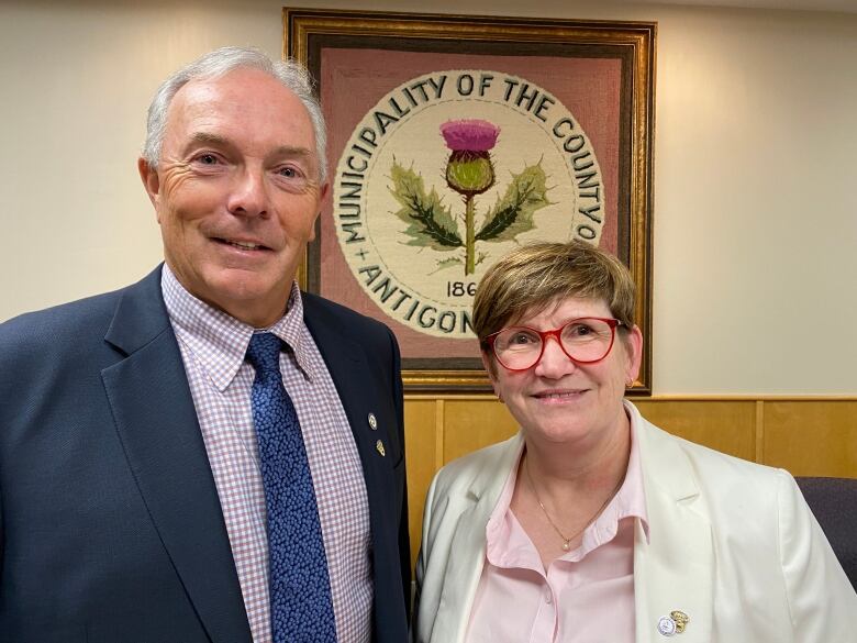 A man and a woman stand together and smile at the camera. A sign in the background says 